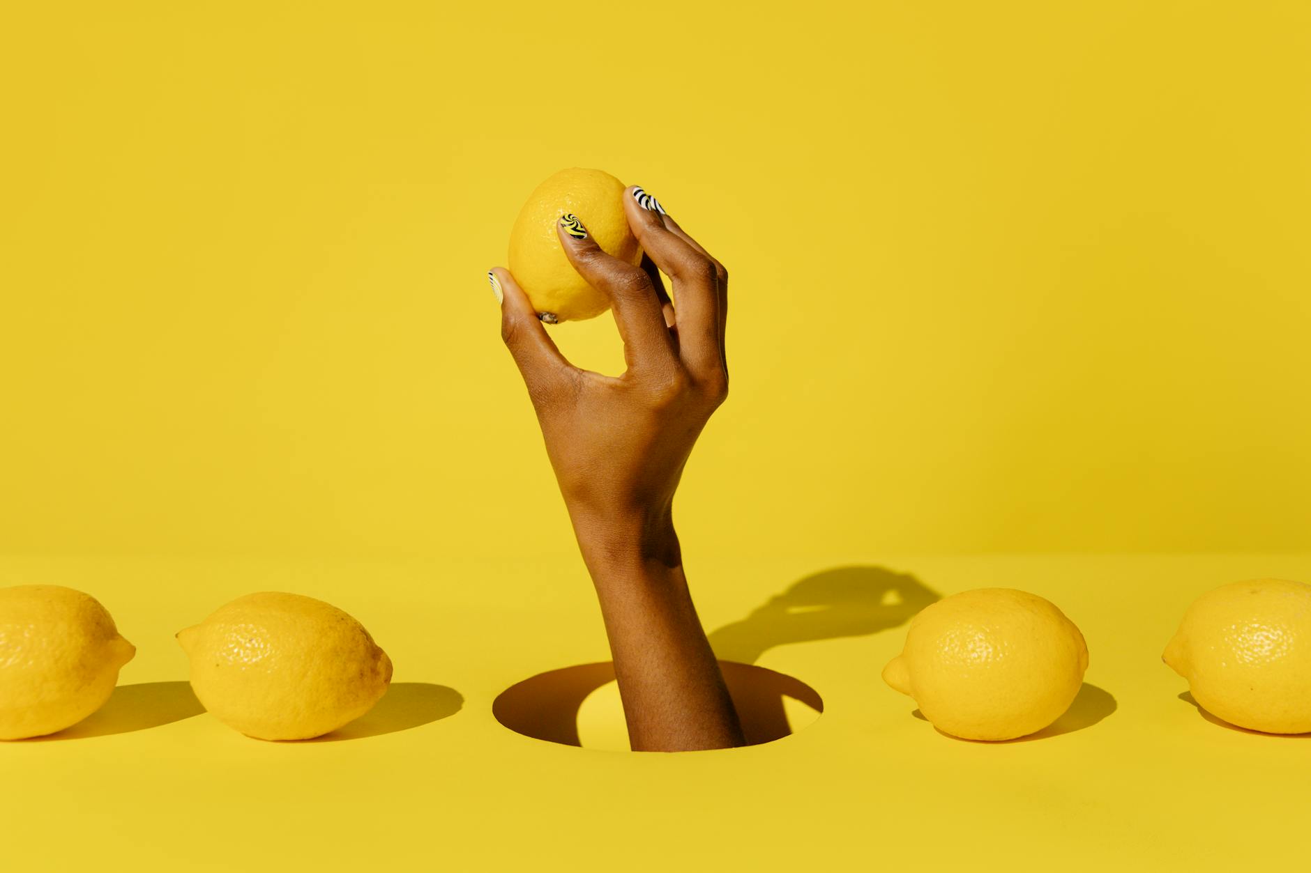 A hand with white nails holding a fresh lemon on a soft pink background, surrounded by additional lemons