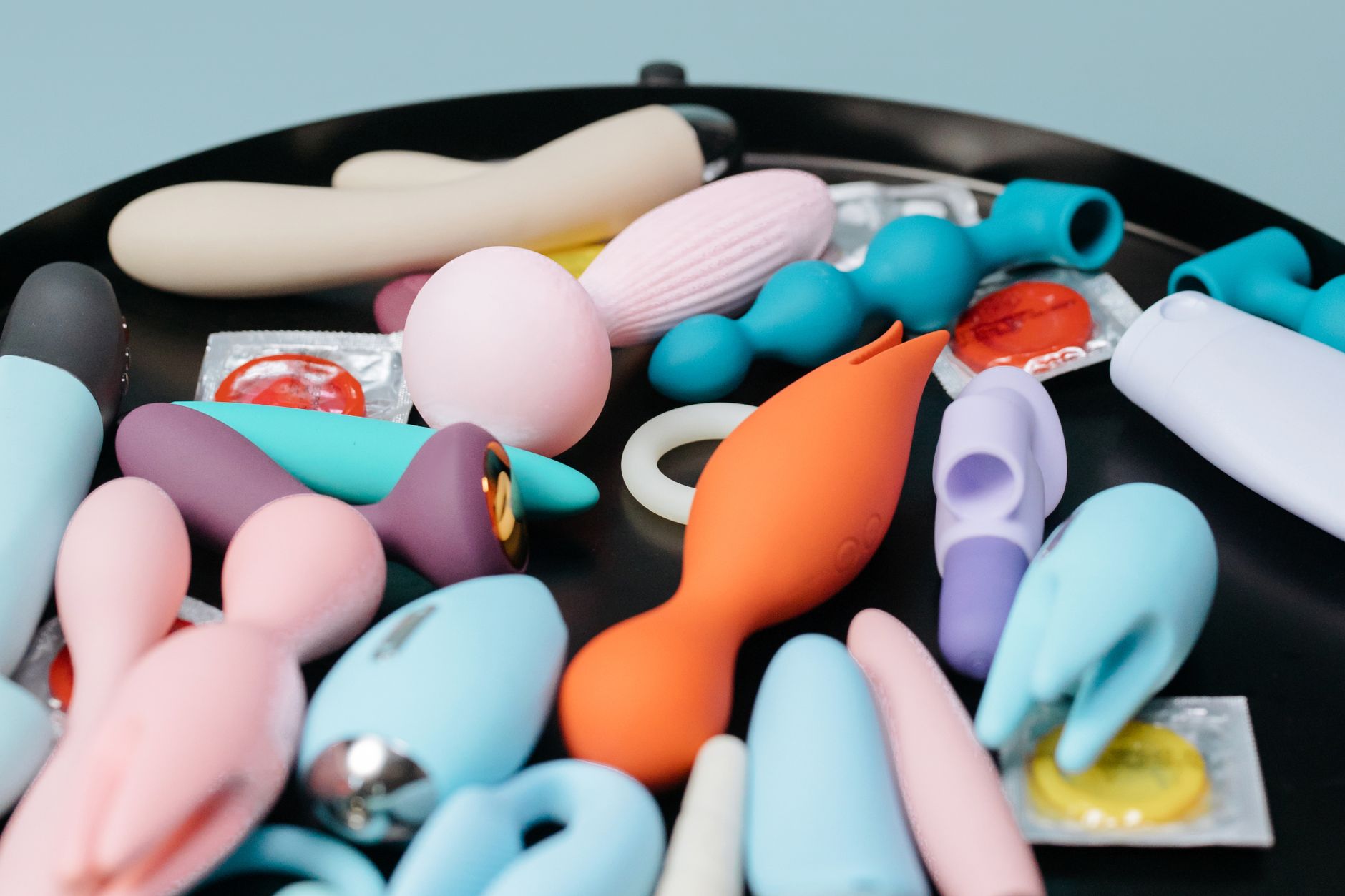 A hand reaching over a variety of colorful sex toys arranged on a table, representing intentional pleasure and self-discovery.